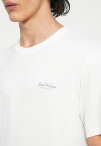 Fehér pamut póló, kerek nyakú és rövid ujjú. A bal mellen kék logó felirat található: "Jack & Jones Archive Selection."
