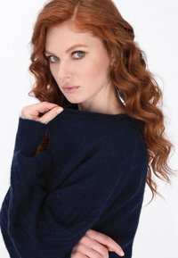 Marineblauer Strickpullover mit lockerer Passform, gerippter Textur und weiten Ärmeln. Das Modell posiert mit der Hand am Ausschnitt und präsentiert den weichen Stoff.