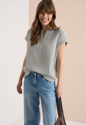 Uśmiechnięta kobieta z długimi brązowymi włosami, ubrana w krótką, paskowaną bluzkę i niebieskie dżinsy, trzymająca pasek do ciemnobrązowej torby.