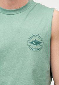 Πράσινο αμάνικο πουκάμισο με κυκλικό λογότυπο Rip Curl Surf Co και κείμενο «Established 69» στο στήθος, φορεμένο από άτομο με ανοιχτόχρωμο δέρμα.