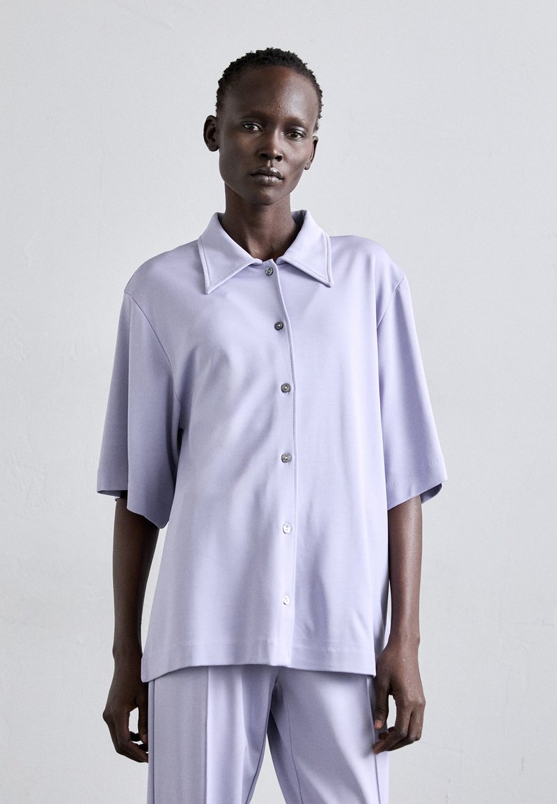 Lavender oversized short-sleeve shirt with a pointed collar, front button closure, smooth fabric texture, and relaxed fit.