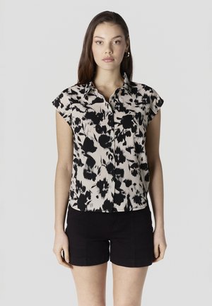 Femeie îmbrăcată cu o cămașă neagră și albă cu nasturi, cu mâneci scurte și imprimeu floral, și pantaloni scurți negri, stând în picioare pe un fundal simplu, deschis la culoare.