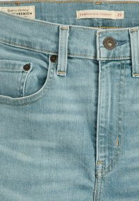 Jean en denim bleu clair en gros plan montrant le bouton avant, les passants de ceinture, les rivets de poche et les étiquettes indiquant la taille 27 et la coupe droite taille haute.