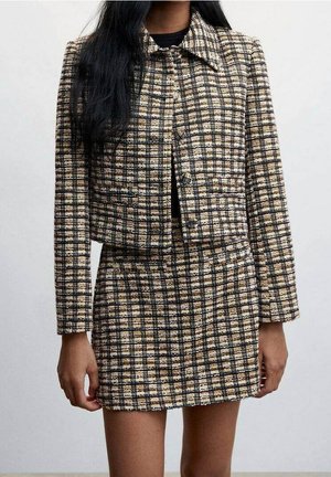 Kobieta ubrana w krótką kurtkę w beżowo-czarno-białą kratę oraz pasującą mini spódniczkę z wysokim stanem na jednolitym tle.