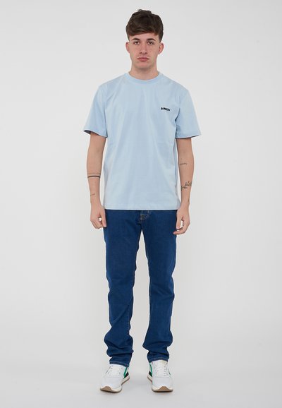 T-shirt azzurro a maniche corte con logo nero, abbinato a jeans blu scuro e sneakers bianche con dettagli verdi. Design semplice e casual.