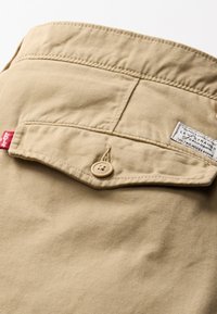 Beige Levi's jackszak met een knoopflap, beige knoop, rode Levi's label en gestikte merklabel nabij de bovenrand.