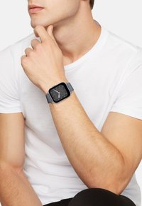Montre connectée avec un cadran noir carré, des aiguilles blanches et un bracelet en silicone gris, portée au poignet d'un teint clair, contre une chemise blanche.