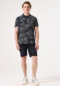Marineblaues Poloshirt mit grünem Blättermuster, kurzen Ärmeln und Kragen, kombiniert mit dunkelblauen Jeansshorts und weißen Sneakers mit grünen Akzenten.