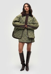 Kvinde iført en oversized, kviltet olivengrøn jakke og matchende nederdel med mørke kanter, parret med knæhøje, mørkebrune hæl-støvler.