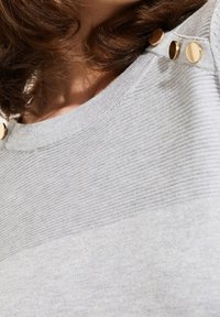 Suéter gris claro hecho de una tela texturizada, con un patrón de canalé y tres botones en tono dorado en el hombro.
