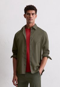 Man in olijfgroene overhemd met knopen over een rood T-shirt, met opgerolde mouwen, staand tegen een effen witte muur.