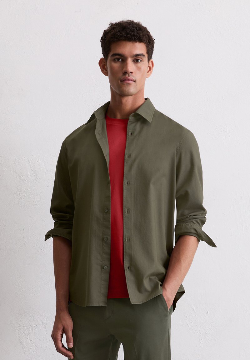Uomo che indossa una camicia verde oliva con bottoni sopra una maglietta rossa, con le maniche arrotolate, in piedi di fronte a un muro bianco semplice.
