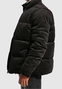 Černá kordurová péřová bunda s vysokým límcem, zapínáním na zip a prošívaným vzorem. Vyznačuje se žebrovanou strukturou a bočními kapsami.