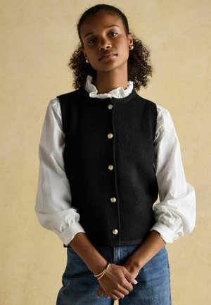 Gilet en laine noir avec des boutons dorés, porté sur une chemise blanche à volants. Le mannequin se tient les bras croisés, les textures du tissu sont visibles.