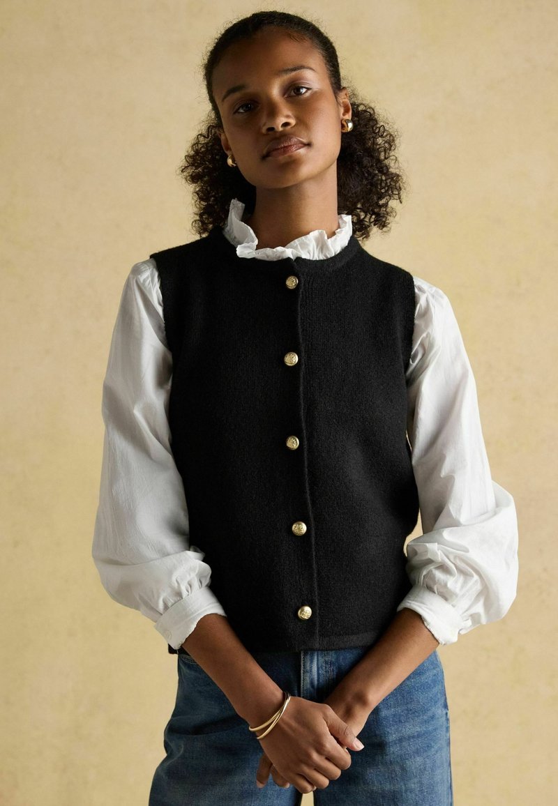 Must must vest, mille kollaste nööpidega, kantud valge volangiga särgi peal. Mudel seisab käed risti, kangatekstuurid on hästi nähtavad.