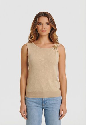 Femme aux cheveux ondulés portant un haut beige sans manches avec un bouton décoratif sur l'épaule gauche et un jean bleu clair, faisant face à l'avant.