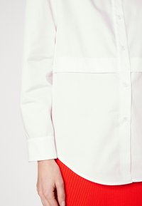 Camicia bianca con colletto, dal tessuto morbido, caratterizzata da una cucitura orizzontale sul petto e maniche lunghe. Abbinata a pantaloni rossi a costine.