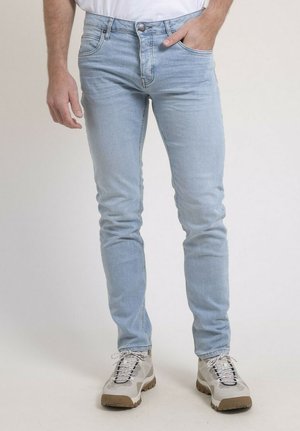 Homme portant un jean slim bleu clair et des baskets blanches, debout avec une main dans la poche sur un fond neutre.