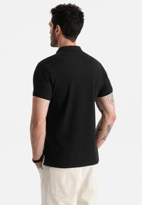 Svart pikétröja med korta ärmar, krage och en skräddarsydd passform. Tyget är slätt, med synliga sömmar vid fållen och ärmarna.