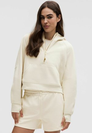 Tejjnys lágy anyagból készült krém színű, rövidített kapucnis pulóver, elülső zsebbel és fél cipzárral, összeillő, gumis derekú rövidnadrággal párosítva.