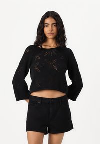 Zwarte gebreide cropped trui met lange mouwen, met een ingewikkeld opengewerkt bloempatroon. Gecombineerd met zwarte denim shorts.