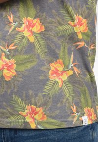Grå stof t-shirt med et farverigt blomstermønster, der indeholder orange blomster og grønne blade. Inkluderer et hvidt mærke i bunden.