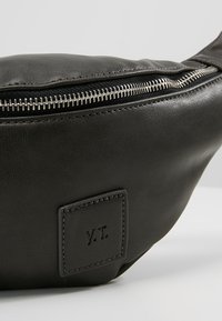 Gros plan sur une pochette en cuir noir avec une fermeture éclair argentée et un patch logo carré cousu avec les initiales "V.T.".
