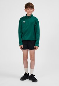 Grøn atletikjakke med lynlås og lange ærmer, sorte shorts, hvide sokker og sorte sneakers. Jakken og shorts har logo.