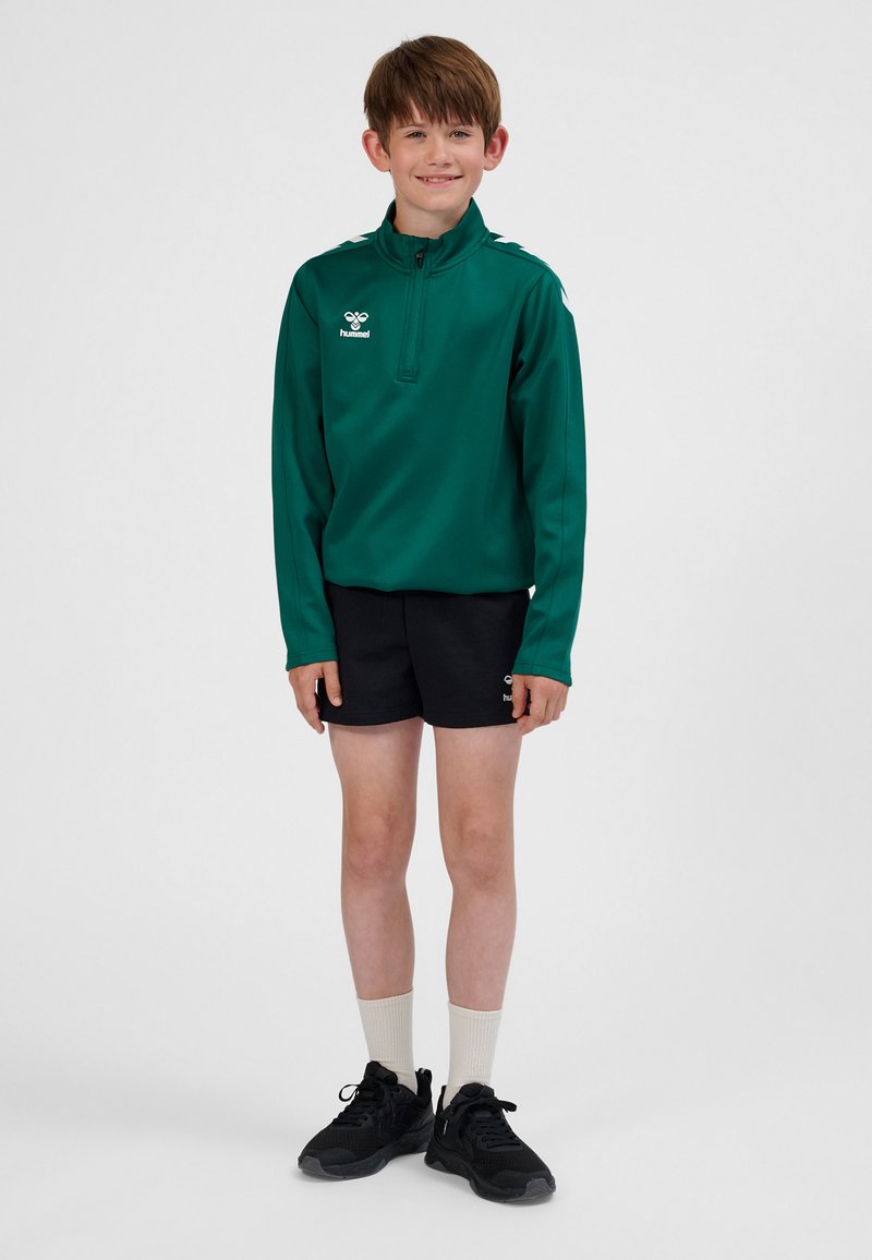 Grøn atletikjakke med lynlås og lange ærmer, sorte shorts, hvide sokker og sorte sneakers. Jakken og shorts har logo.