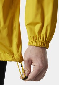 Chaqueta amarilla ligera con puños elásticos, dobladillo con cordón y un acabado suave y texturizado. La manga muestra una mano ajustando el dobladillo.