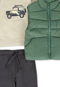 Gilet verde imbottito con zip e tasche, camicia beige con grafica di un veicolo e pantaloni sportivi neri con coulisse. Materiali testurizzati e morbidi.