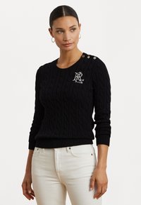 Zwarte kabelgebreide trui met geribde rand, voorzien van decoratieve knopen op de schouder en geborduurd logo op de borst. Gecombineerd met witte jeans.