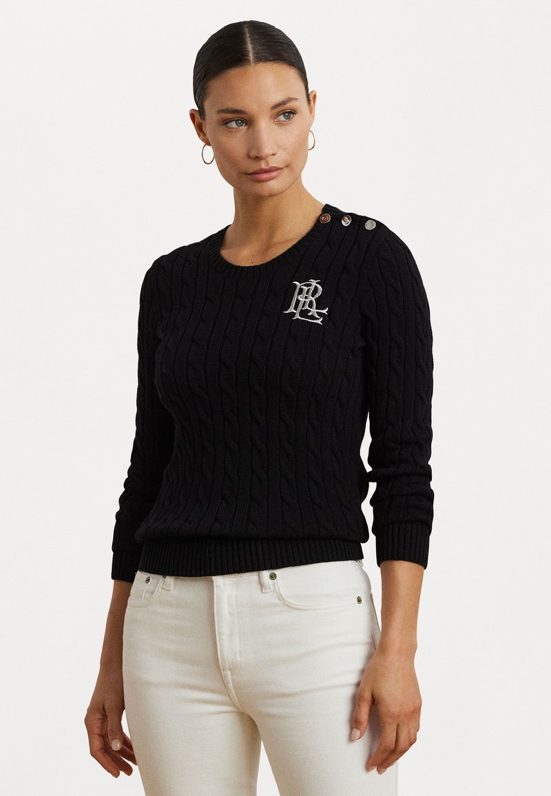 Zwarte kabelgebreide trui met geribde rand, voorzien van decoratieve knopen op de schouder en geborduurd logo op de borst. Gecombineerd met witte jeans.