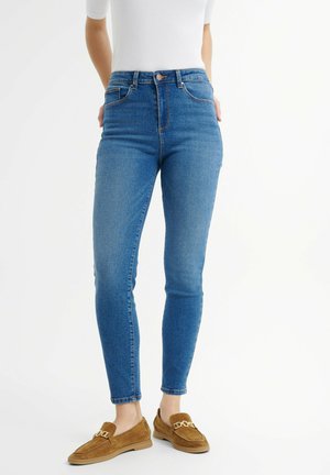 Niebieskie jeansy typu skinny z wysokim stanem, gładką teksturą denimu i pięcioma kieszeniami. Zestawione z brązowymi mokasynami z dekoracyjnym łańcuchem.
