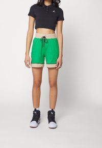 Černé zkrácené polo s bílým logem, v kombinaci se zelenými a béžovými šortkami na šňůrku a černými vysokými teniskami s bílou podrážkou.