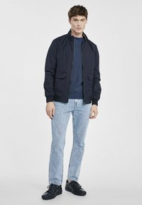 Granatowa kurtka bombowiec z zamkiem z przodu, z dwiema kieszeniami z przodu, noszona na teksturowanym niebieskim swetrze, zestawiona z jasnymi dżinsami i czarnymi trampkami.