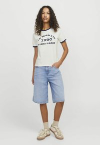 Crème t-shirt met korte mouwen en marineblauwe rand en bedrukte tekst. Draagt samen met lichtblauwe spijkerbroeken en beige sneakers met subtiele accenten.