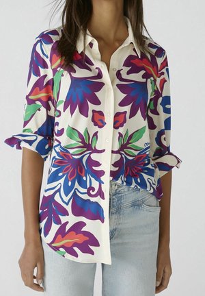 Femme portant une chemise blanche à boutons avec de grands motifs floraux colorés en violet, bleu, rouge et vert, associée à un jean bleu clair.