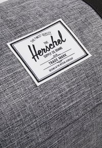 Šedá látková taška s bílou etiketou a černým textem "Herschel Supply Co. Brand" a dekorativními prvky. Texturovaný povrch s tkaným vzhledem.