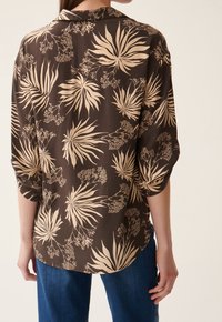 Chemise boutonnée marron avec un motif floral crème. Coupe ample, manches trois-quarts, ourlet arrondi et texture de tissu lisse.