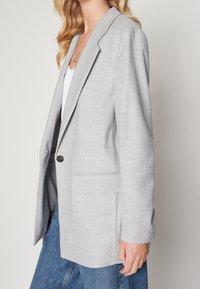Blazer gris clair en tissu lisse, avec une fermeture à un seul bouton, deux poches avant et une coupe légèrement oversize.