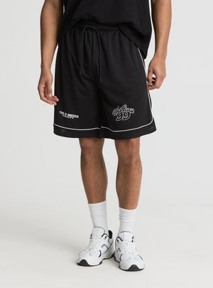 Uomo che indossa pantaloncini neri in rete con bordi bianchi e scritte "This is America" e "Wax Coax 99", abbinati a calze bianche e sneakers.