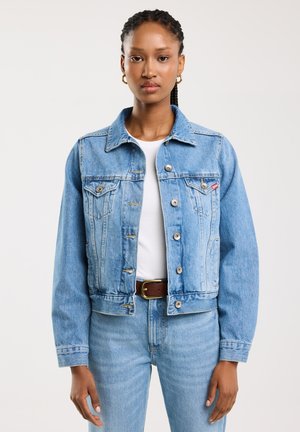 Veste en denim bleu clair avec une coupe classique, dotée de deux poches poitrine et de boutons en argent, assortie à un haut blanc et un jean bleu.