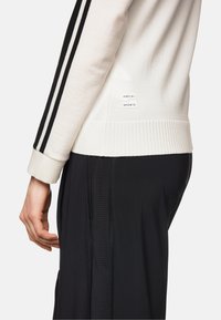 Weißes Sweatshirt mit schwarzen Streifen und geripptem Saum, kombiniert mit schwarzen strukturierten Hosen. Verfügt über ein Logotag an der Seite des Sweatshirts.
