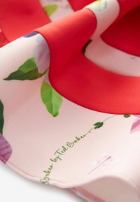 Tissu doux avec une bordure rouge et un imprimé floral, comprenant des feuilles vertes et une signature « Baker by Ted Baker » sur un fond rose pâle.