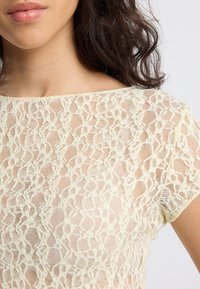 Close-up of a woman wearing a pale yellow lace top with a floral pattern and short sleeves, showing her lips and dark wavy hair.