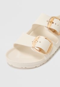 Sandali bianchi con due ampi strap, dotati di minuteria in tono oro. Suola testurizzata per una migliore aderenza, realizzati in materiale liscio e flessibile.