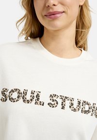 Cremfarbener Rundhalsausschnitt-Pullover mit großem Text "SOUL STUDIO" im Leopardenmuster, aus weichem Baumwollmaterial.
