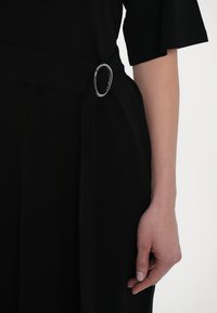 Crna odjeća s širokim pojasom koji ima okruglu srebrnu kopču. Mekana, teksturirana tkanina elegantno pada. Ruka modela naslonjena je sa strane.