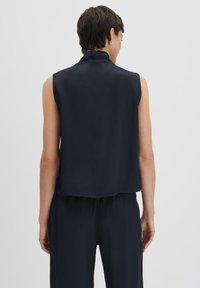 Top senza maniche di colore blu navy con colletto alto, texture liscia e design fluido. Orlo leggermente curvo, abbinato a pantaloni in tessuto coordinato.